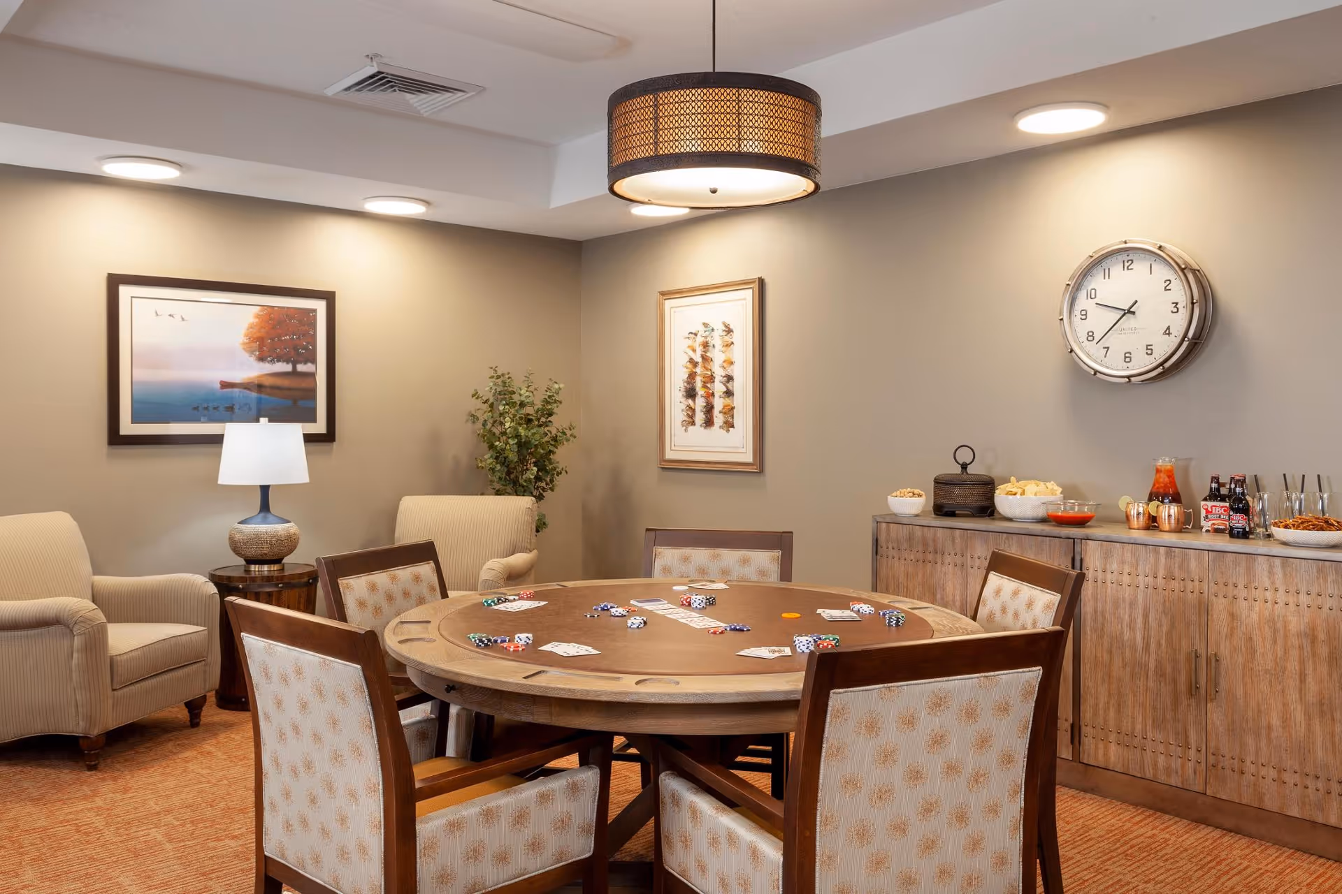 A cozy interior room with a round poker table surrounded by six cushioned chairs. The table has playing cards and poker chips scattered on it. In the background, there are two beige armchairs with a small round table and lamp between them, a plant, framed artwork on the walls, a large wall clock, and a wooden cabinet topped with snacks and drinks.