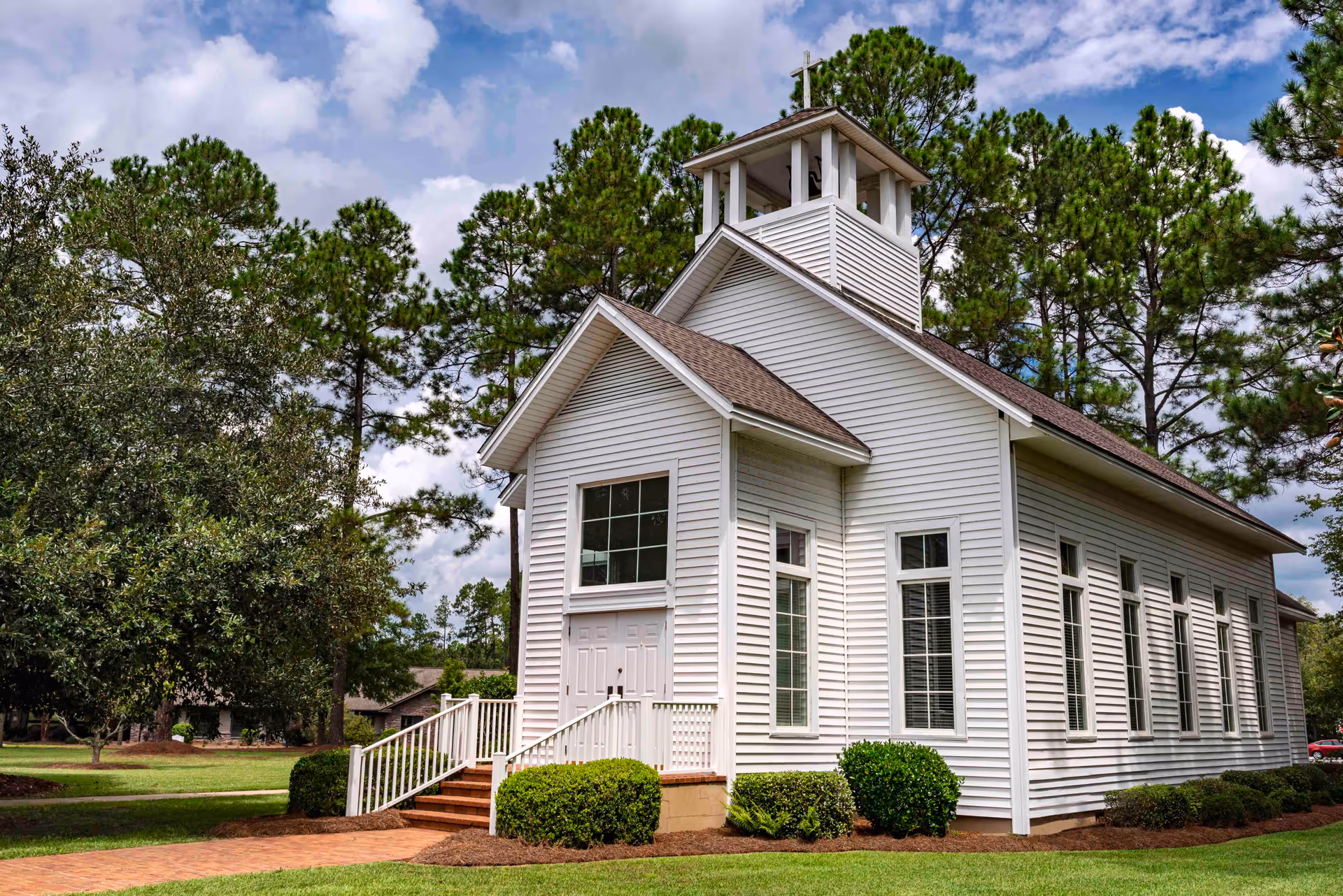 A white wooden chapel-style building with a small bell tower and cross on top, surrounded by green trees and bushes under a partly cloudy sky.
