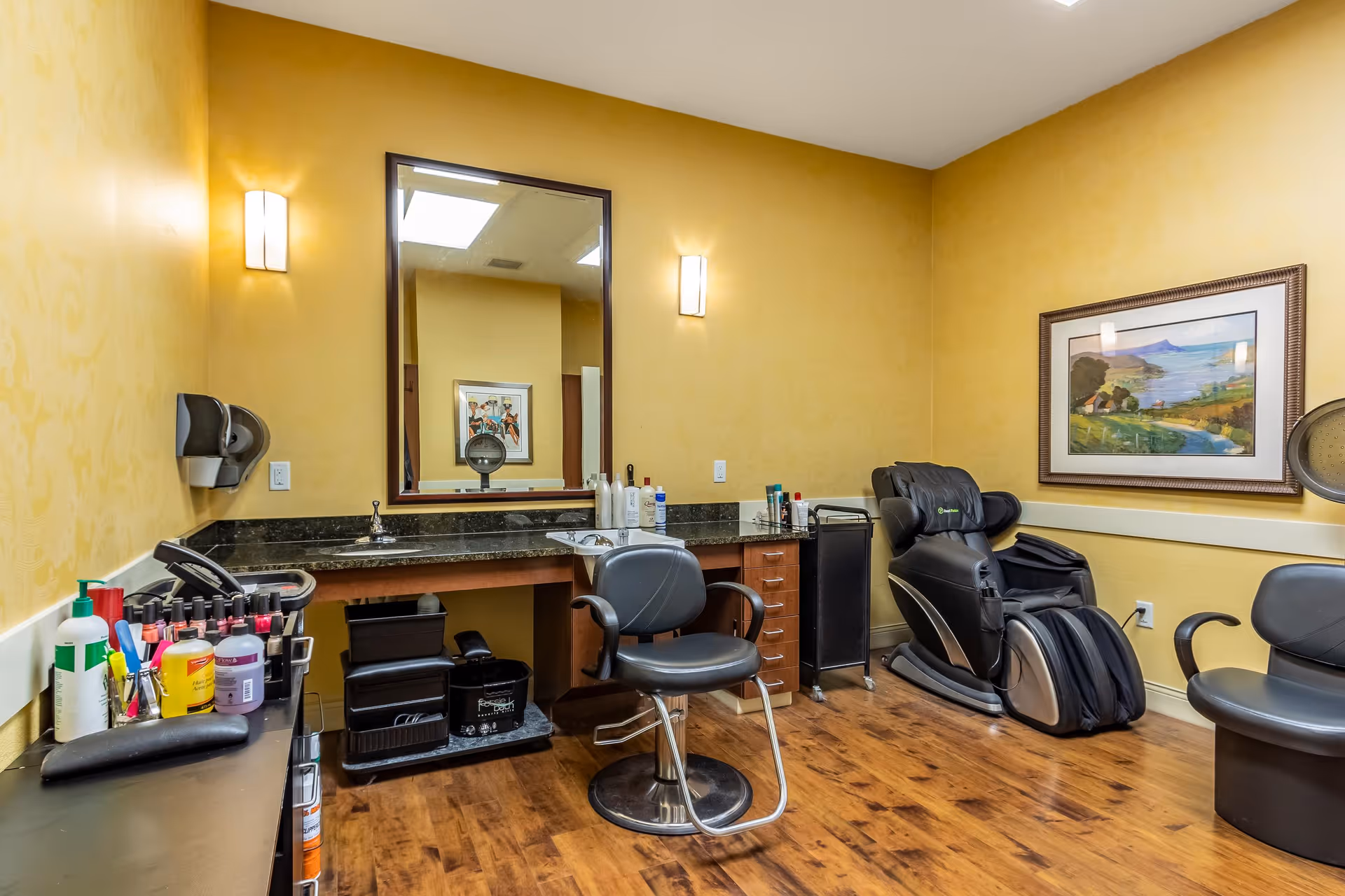 A salon room with yellow walls and wooden flooring featuring a black salon chair in front of a large mirror above a countertop with various hair care products. To the right, there is a black massage chair and another black chair. A framed landscape painting hangs on the wall.