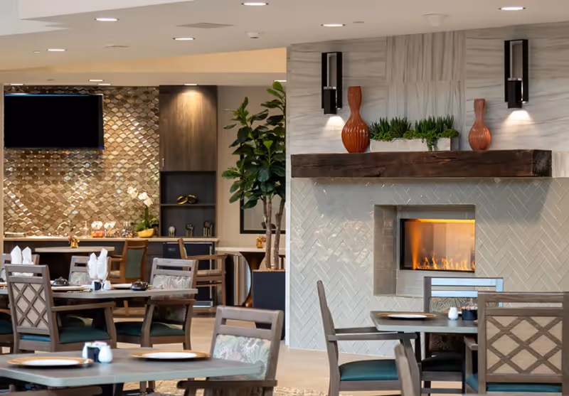 A cozy dining area in a senior living facility featuring wooden tables and chairs with patterned cushions. There is a modern fireplace with a wooden mantel decorated with two orange vases and green plants. The background includes a wall with a decorative tile backsplash, a mounted TV, and additional seating and cabinetry.