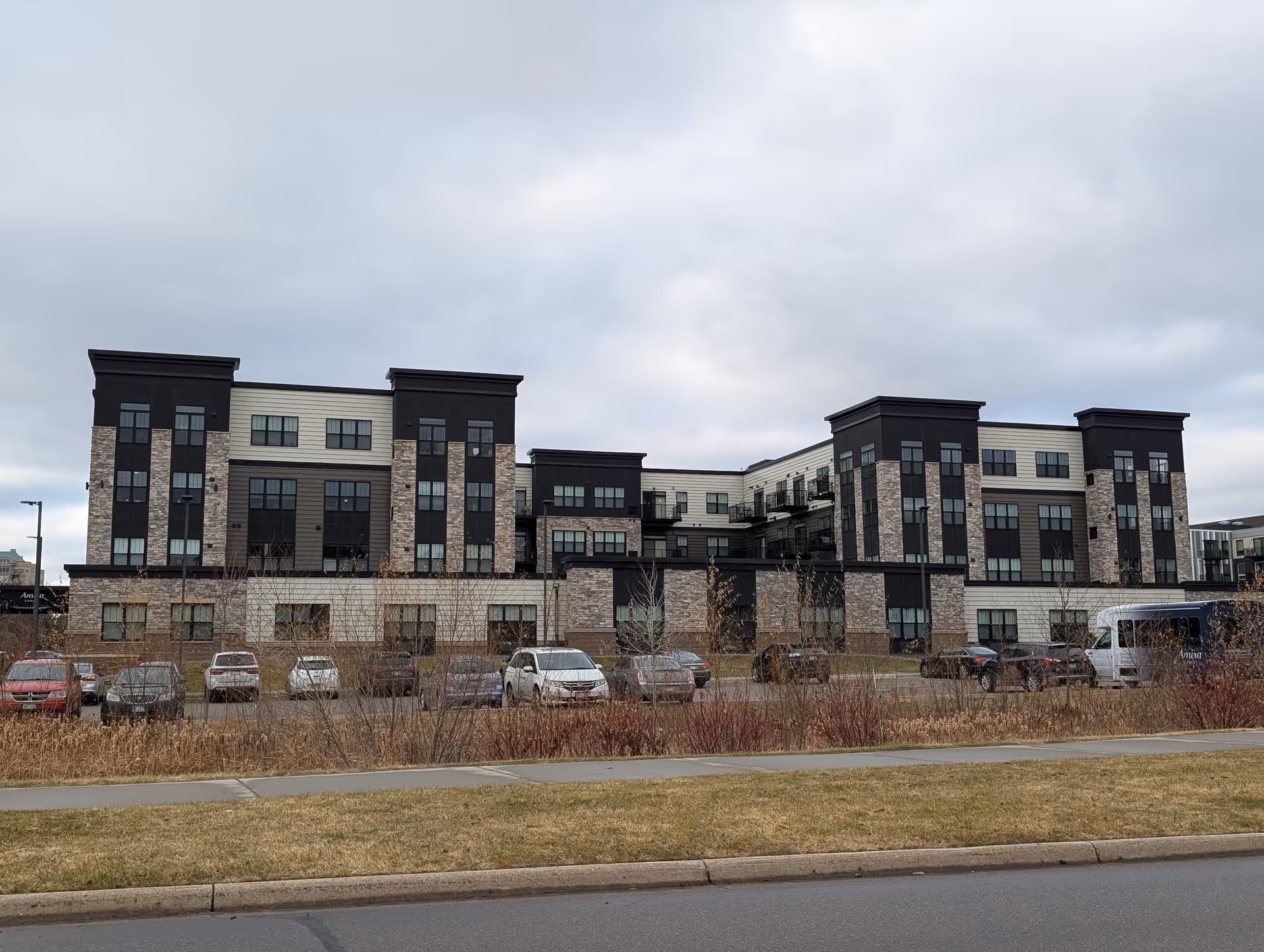 Exterior view of a modern multi-story building with a combination of dark and light siding and stone accents. Several cars are parked in front of the building, and there is a grassy area with a sidewalk in the foreground under a cloudy sky.