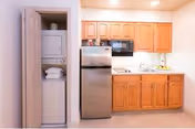 Small kitchenette with wooden cabinets, a stainless steel refrigerator and microwave, a sink and counter, and a stacked washer-dryer in an open closet.