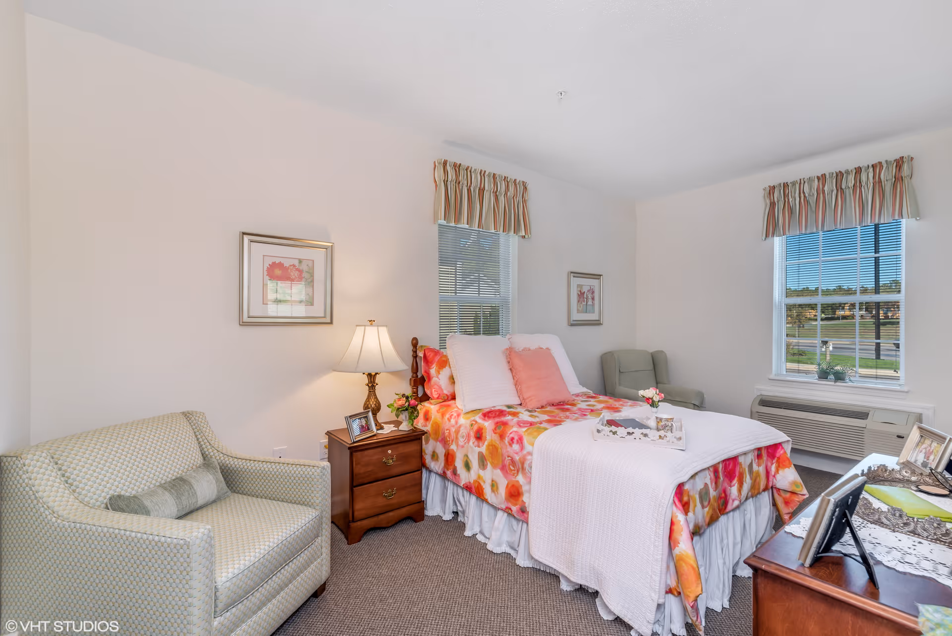 Bright furnished bedroom with a floral-print bed, nightstand and lamp, two armchairs, and windows with valances.