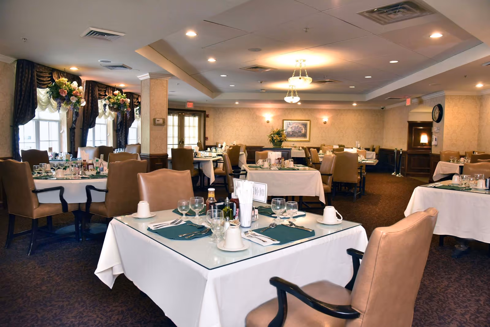 A spacious dining room with multiple tables covered in white tablecloths and set with glassware, plates, and utensils. The room features beige walls, carpeted floors, large windows with dark curtains and floral arrangements, and warm ceiling lighting. Comfortable padded chairs surround each table, creating an inviting atmosphere.
