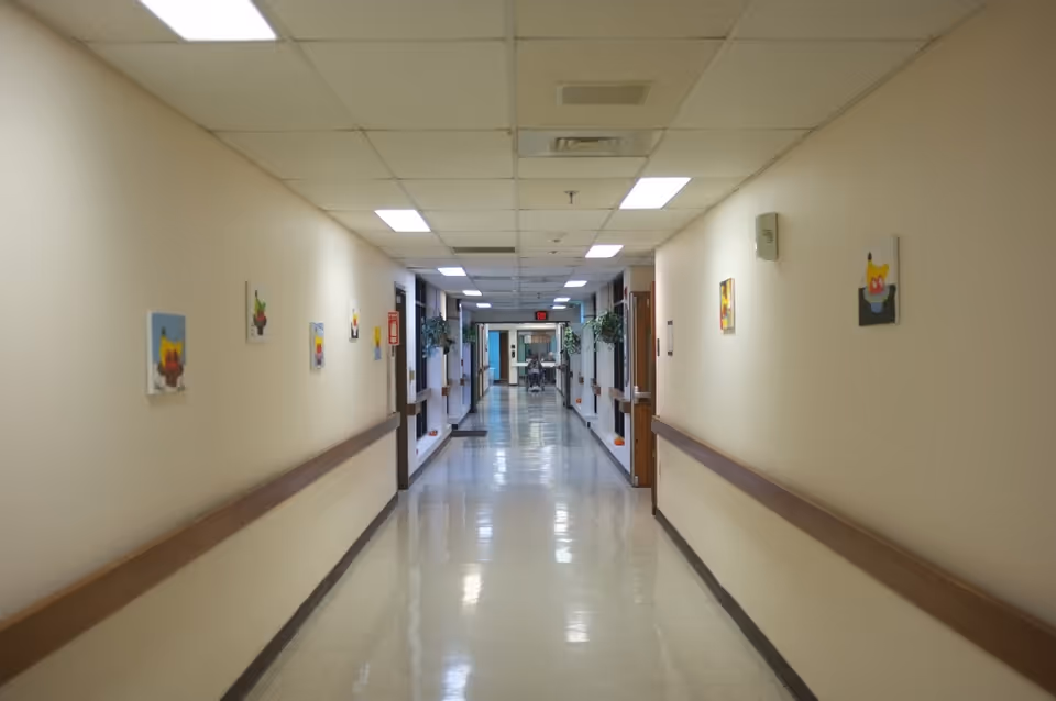 A long, clean hallway in an assisted living facility with beige walls, handrails on both sides, small colorful paintings hanging on the walls, and bright overhead fluorescent lighting. At the far end, a person in a wheelchair is visible near the exit door.
