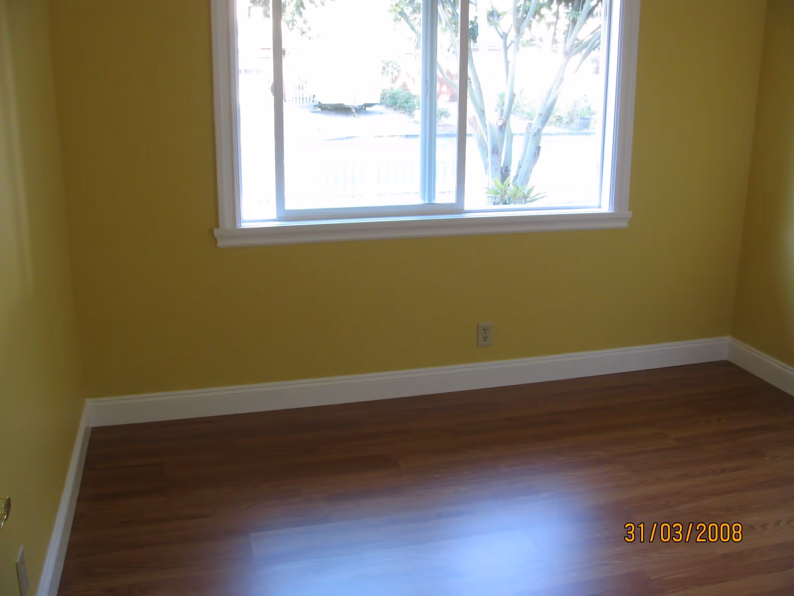 Empty room with yellow walls, a large window showing trees outside, and wooden flooring.