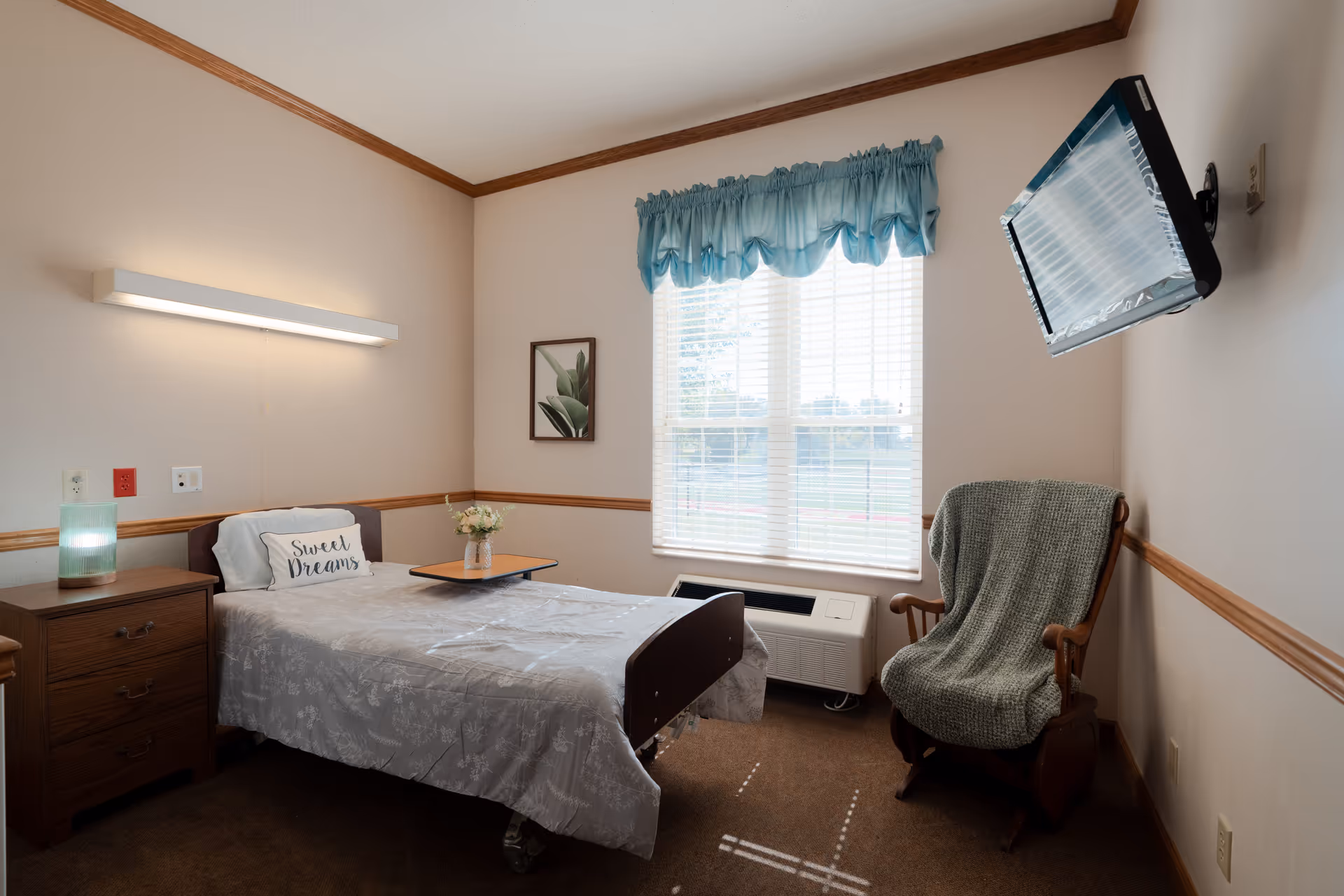 A small, cozy nursing facility bedroom with a single bed covered in a light gray floral bedspread and a pillow that says 'Sweet Dreams'. Next to the bed is a wooden nightstand with a lamp and a small vase of flowers on a tray table. A window with blue valance curtains lets in natural light. A wooden rocking chair with a green knitted blanket is placed near the window, and a flat-screen TV is mounted on the wall opposite the bed.
