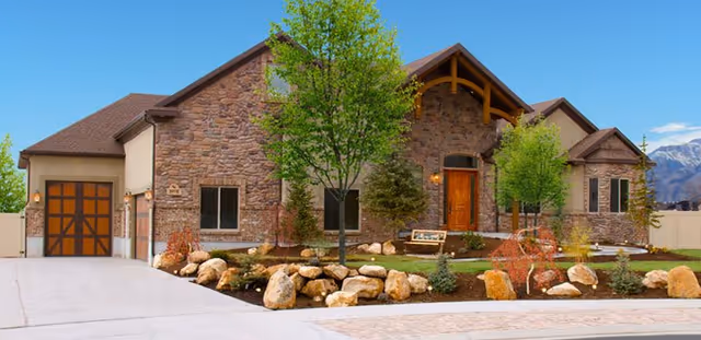 Front exterior view of a single-story assisted living facility building with stone facade, a wooden front door, large windows, a garage, landscaped garden with rocks and trees, and mountains in the background under a clear blue sky.