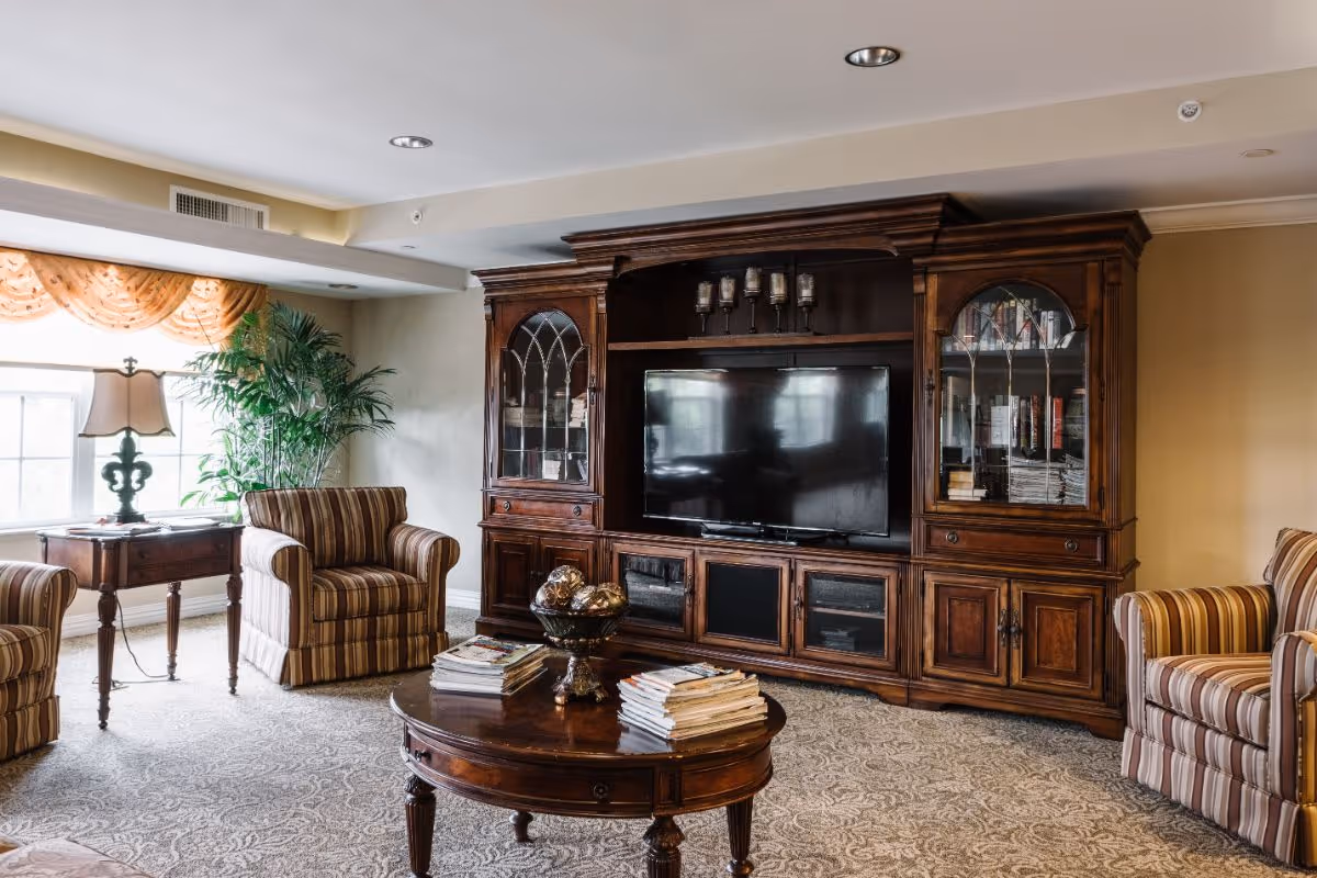 Cozy living room with a large wooden entertainment center and TV, striped armchairs, a coffee table with books, and a window with drapes.