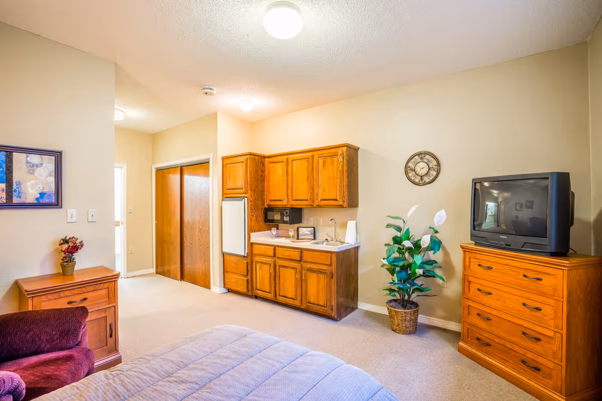 Interior view of a senior living facility room featuring a bed with a light blue quilt, a wooden nightstand with a small flower pot, a maroon armchair, a kitchenette with wooden cabinets, a microwave, and a small sink. There is a wooden dresser with an old-style television on top, a potted plant beside it, and a wall clock above the plant. The walls are painted beige, and there is a framed picture on one wall.