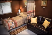 A cozy room with a single bed covered with a patterned quilt, a bedside table with a lamp, a window with blinds, and a dark brown leather couch with two yellow pillows. The walls are painted brown and there is a curtain partially drawn near the bed.