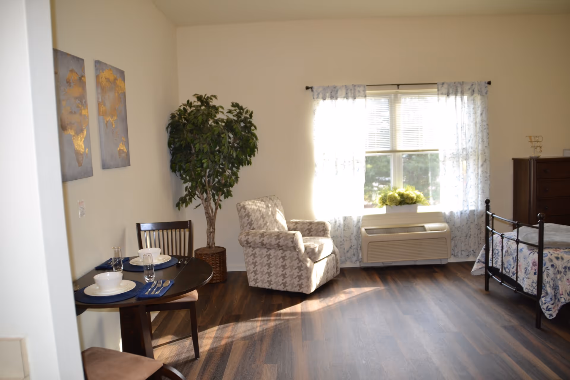 Sunlit studio-style bedroom with an armchair, small dining table, bed, dresser and a window air conditioning unit.
