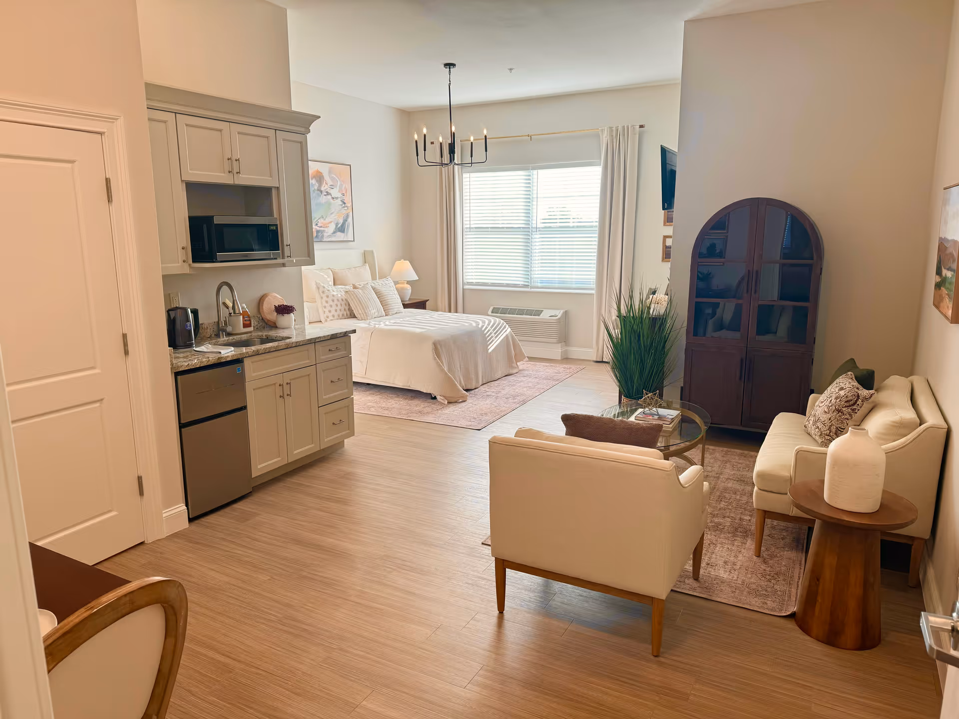 A bright and spacious assisted living studio apartment featuring a small kitchenette with cabinets, a microwave, mini fridge, and sink on the left. In the background, there is a neatly made bed with pillows and a bedside lamp. To the right, there is a cozy living area with a sofa, armchair, coffee table, side table with a decorative vase, and a wooden cabinet. A large window with blinds and curtains lets in natural light, and the floor is covered with light wood laminate.