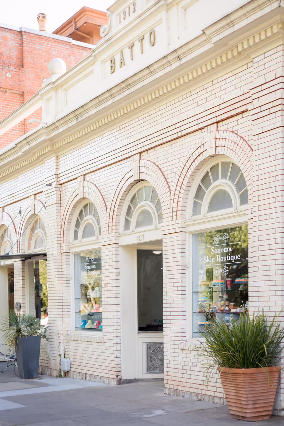 Exterior view of a light-colored brick building with arched windows and a door in the center. The windows display signs that read 'Sonoma Skin Boutique.' There are two large potted plants on either side of the entrance and a bench to the left.