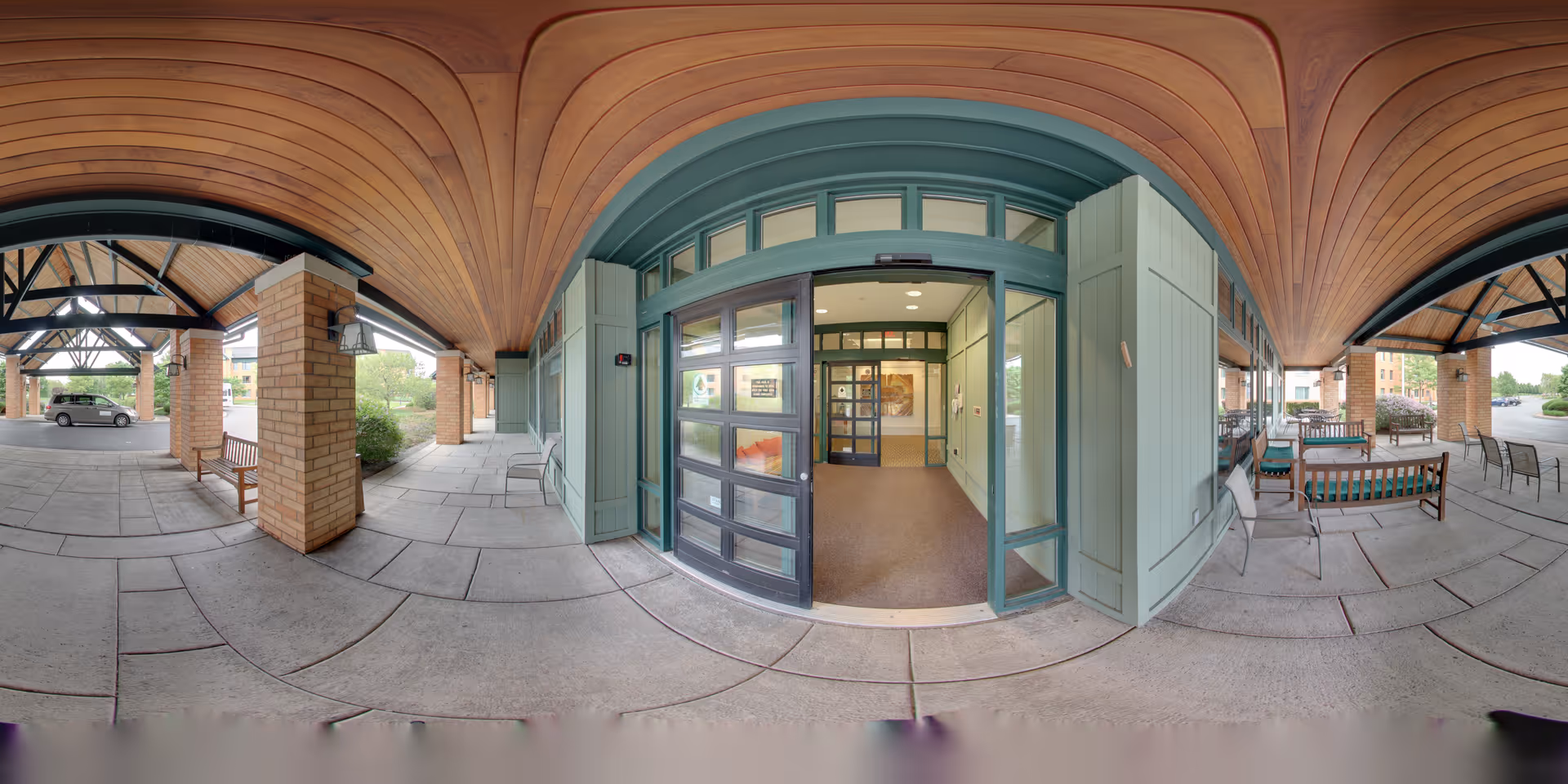 Covered entrance area of The Summit at Brighton Independent Living facility with a wooden arched ceiling, brick pillars, benches, and glass doors leading inside. Outdoor seating is visible on the right side under the covered area.