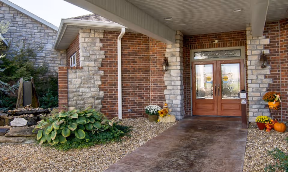 Entrance to a brick and stone building with double wooden doors featuring decorative glass panels. The covered walkway is surrounded by gravel landscaping with green plants and autumn-themed decorations including pumpkins and flowers.