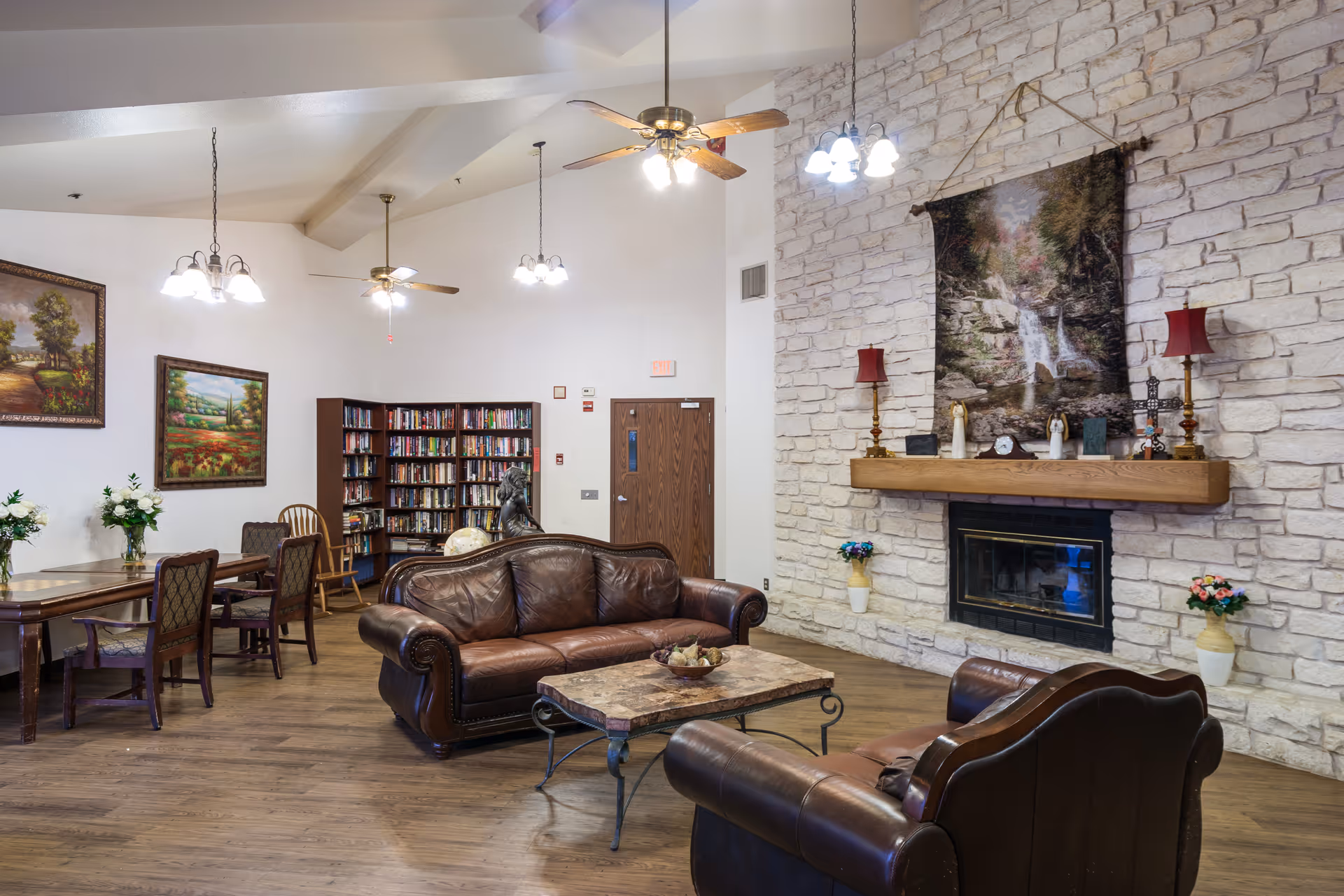 A cozy living room area in a senior living facility with two brown leather sofas, a stone fireplace with a wooden mantle, decorative lamps, and a large tapestry above the fireplace. There is a wooden coffee table in front of the sofas, a bookshelf filled with books, a wooden dining table with chairs, and paintings on the walls. The room has wooden flooring and ceiling fans with lights.