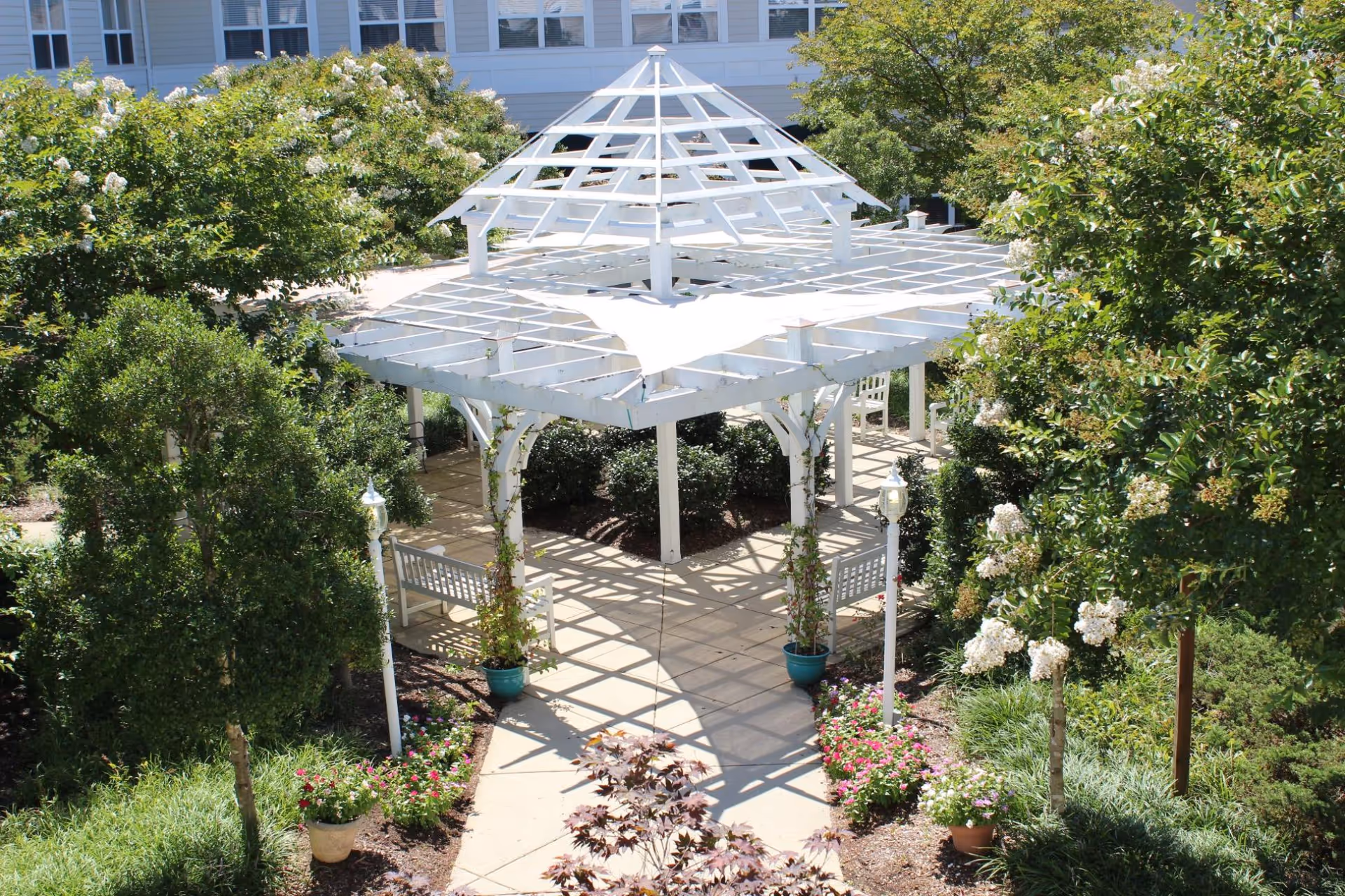 White wooden pergola-style gazebo in a landscaped courtyard with benches and flowering shrubs in front of a building.