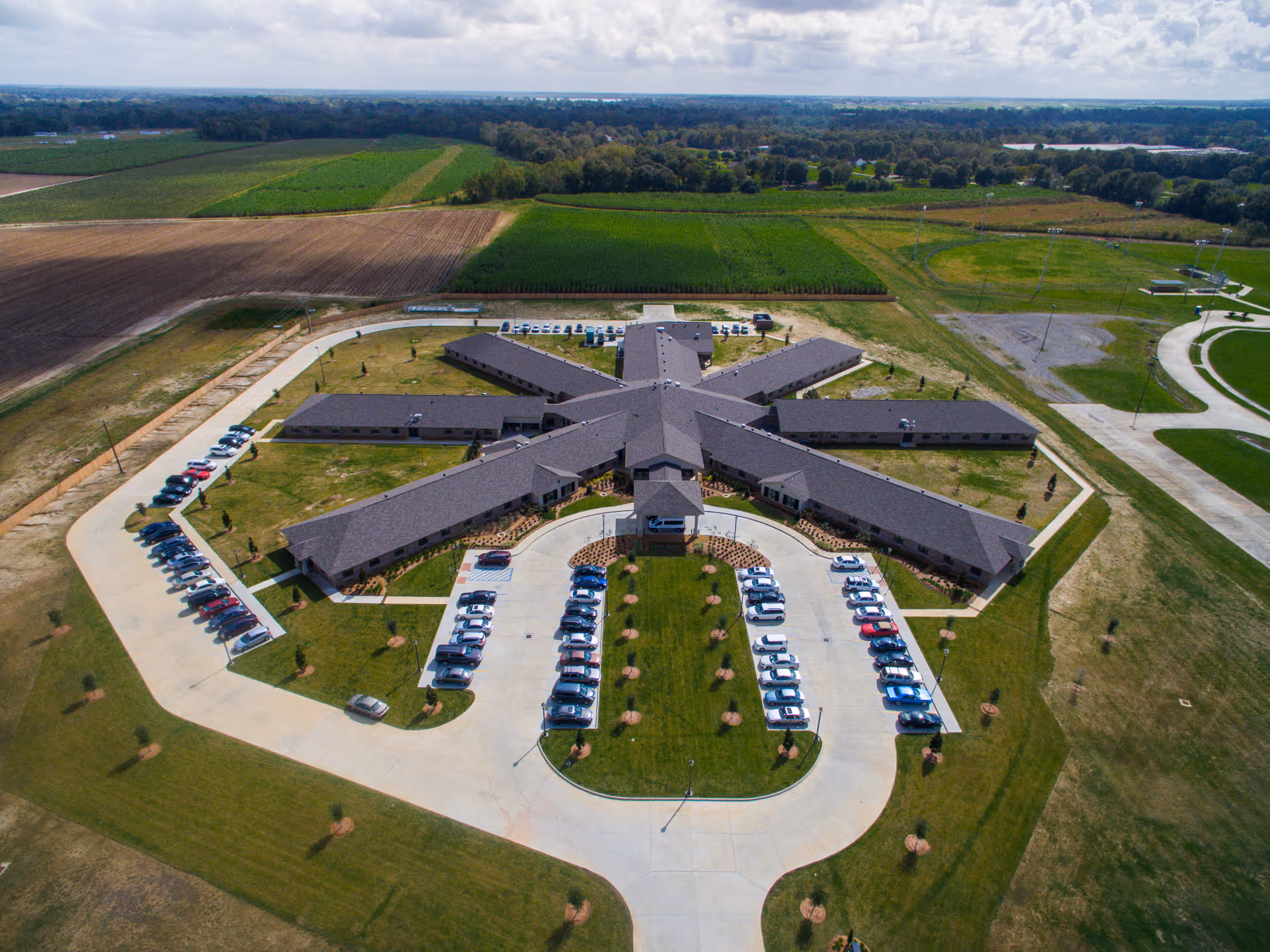 Aerial view of a star-shaped senior living facility with radiating wings, a central entrance, surrounding parking lots and open fields.