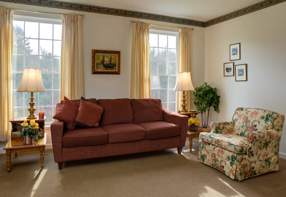 A cozy living room with a brown sofa and a floral armchair. Two large windows with cream-colored curtains let in natural light. There are two side tables with lamps and decorative plants, and framed artwork on the walls.