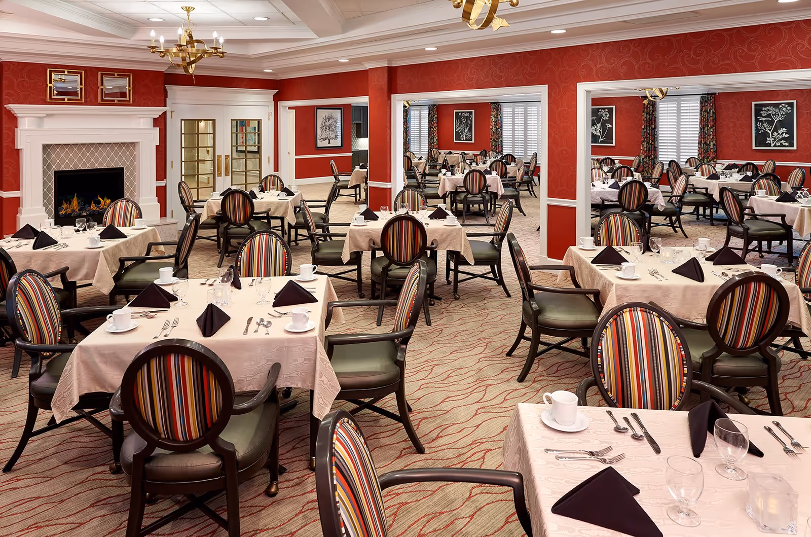 A spacious dining room with multiple tables covered in beige tablecloths, each set with black folded napkins, white cups, glasses, and silverware. The chairs have dark wooden frames with striped upholstery in red, orange, yellow, and black. The walls are painted red with white trim, and there is a white fireplace with a lit fire on the left side. The room is well-lit with ceiling lights and two gold chandeliers. There are framed botanical prints on the walls and large windows with floral curtains.
