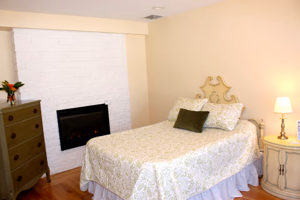 Bedroom with a patterned bedspread on a double bed, decorative headboard, wall-mounted fireplace, dresser and bedside table with a lamp.