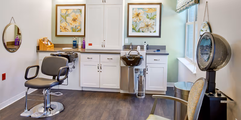 A bright salon-style room with styling chairs, shampoo sinks, cabinets, and a hooded hair dryer by a window.