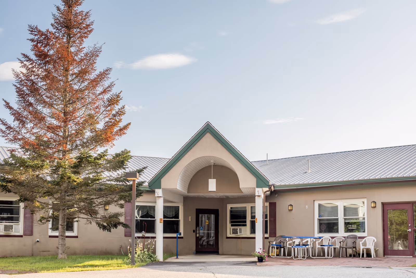 Front entrance of a single-story senior living facility with a covered portico, outdoor chairs, and a large tree nearby.