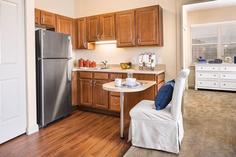 A compact kitchen area with wooden cabinets, a stainless steel refrigerator, and a small countertop table set with a cup, bowl, and placemat. A white chair with a blue cushion is positioned at the table. Adjacent to the kitchen is a carpeted room with a white dresser and window blinds.