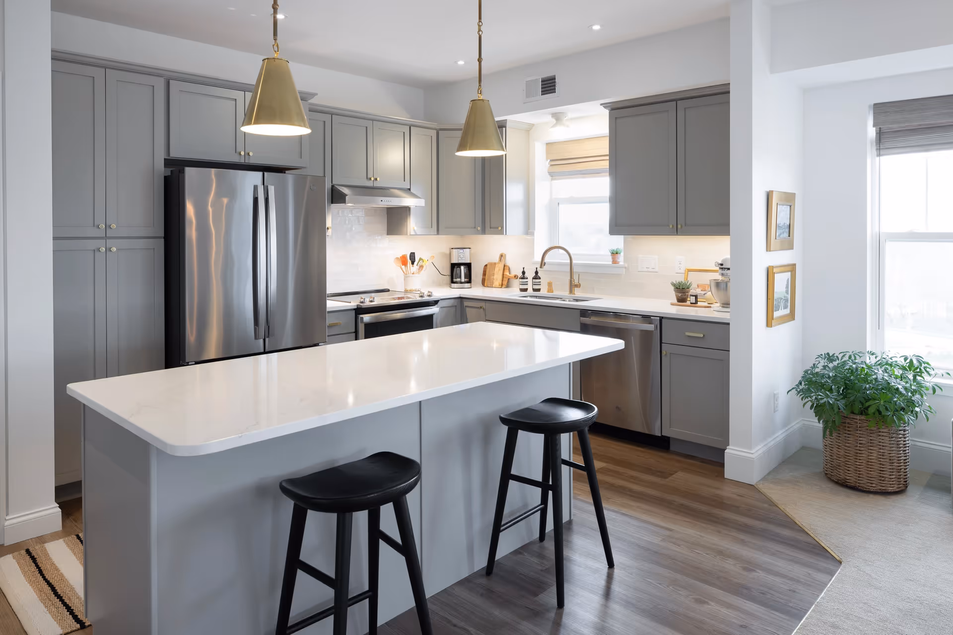 Modern kitchen with gray cabinets, stainless steel refrigerator, oven, and dishwasher. A white island countertop with two black stools is in the center. Two pendant lights hang above the island. There is a window with a beige shade, a potted plant in a woven basket near the window, and framed pictures on the wall.