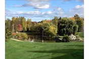 A scenic outdoor view featuring a small pond surrounded by lush green grass, trees with autumn foliage, and a partly cloudy blue sky.
