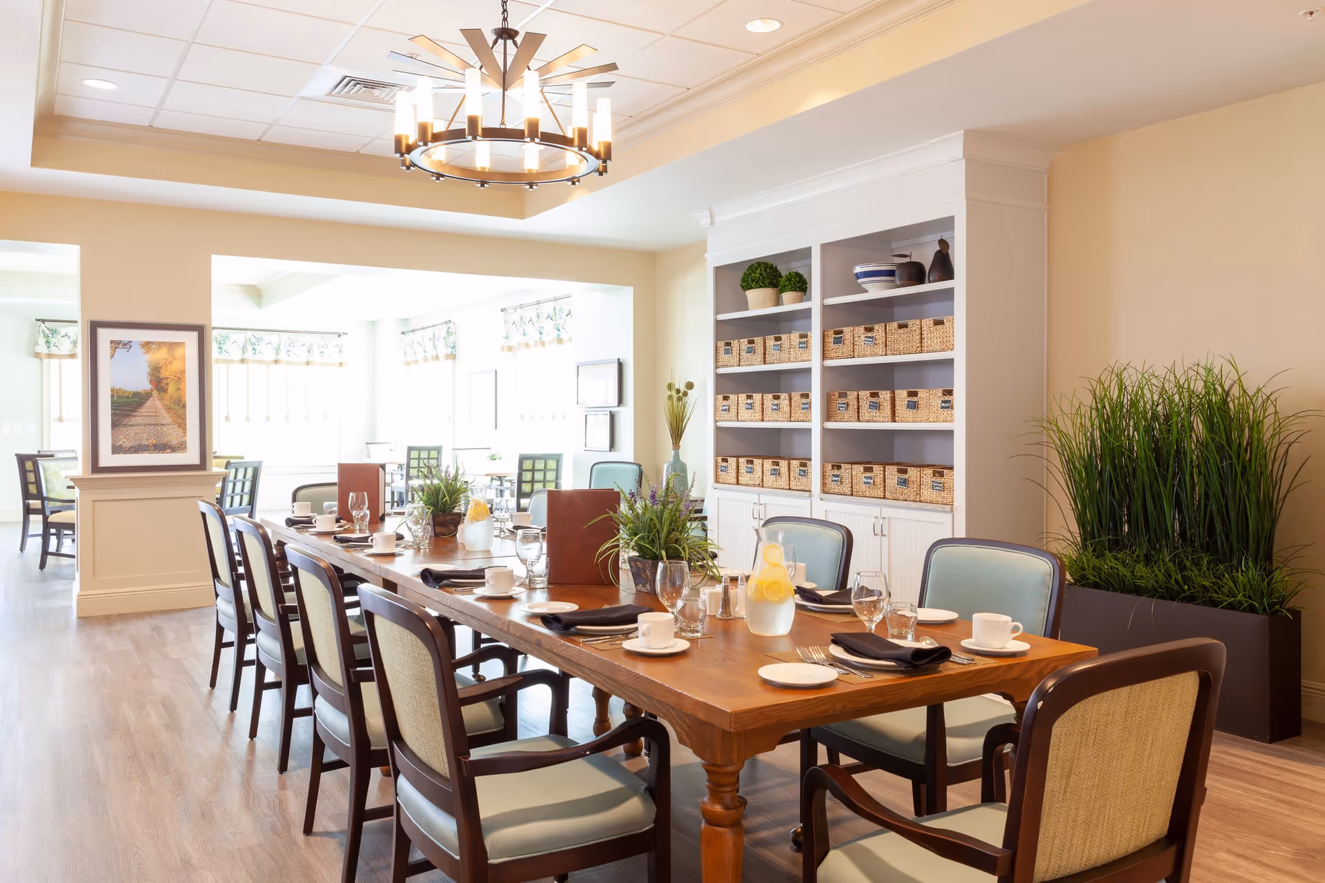 A bright and spacious dining room with a long wooden table set with cups, plates, glasses, and black napkins. The room features light-colored walls, a large white shelving unit with wicker baskets, green plants, and a modern chandelier hanging from the ceiling. Additional tables and chairs are visible in the background near large windows with floral valances.