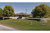 Single-story residential care building with a circular driveway, lawn, trees, and a stone entrance sign.
