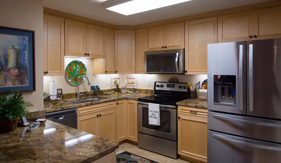 A modern kitchen with light wood cabinets, granite countertops, and stainless steel appliances including a refrigerator, stove, microwave, and dishwasher. The kitchen features a double sink, decorative plates on the wall, framed artwork, and a towel hanging on the oven door that reads 'Salt Water Heals Everything.'
