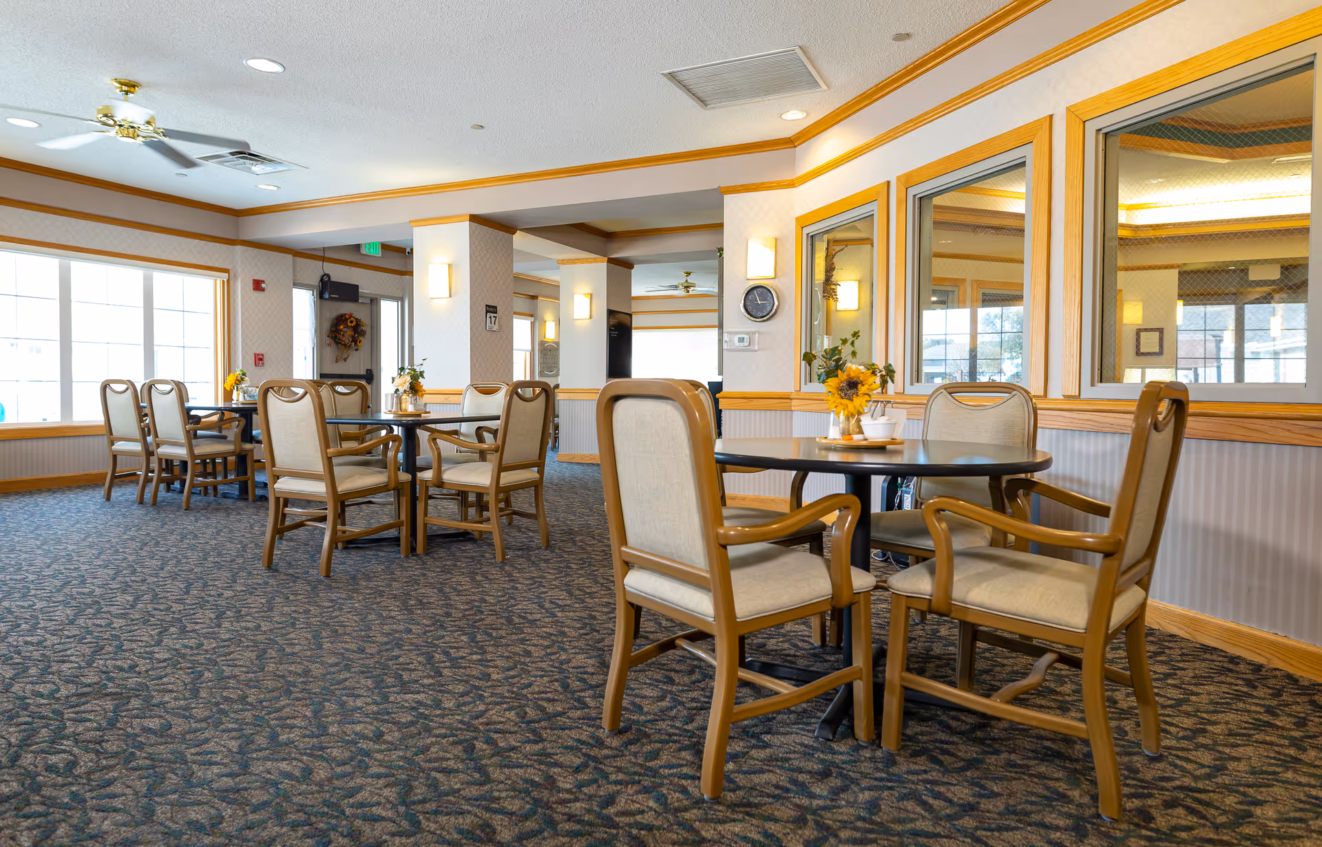 A bright and spacious dining area in an assisted living facility with multiple round tables surrounded by beige cushioned chairs. The room features large windows allowing natural light, wall mirrors, and floral centerpieces on each table. The carpet has a patterned design, and the walls are decorated with light-colored wallpaper and wooden trim.