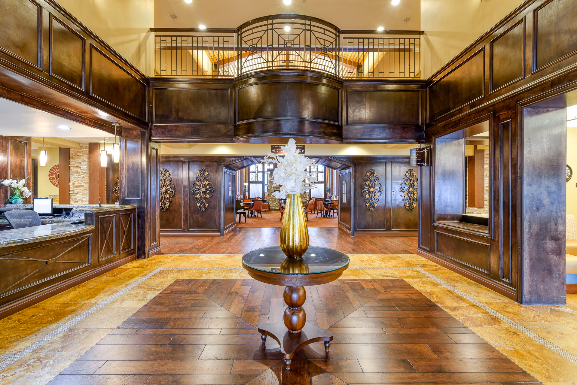 Spacious elegant lobby with a central round table and vase, dark wood paneling, reception desk, and seating area beyond.