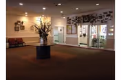 Interior view of a nursing facility lobby with a round table in the center holding a large floral arrangement. There are chairs along the left wall, bulletin boards, and glass doors leading to another area. The walls are decorated with photos and notices.