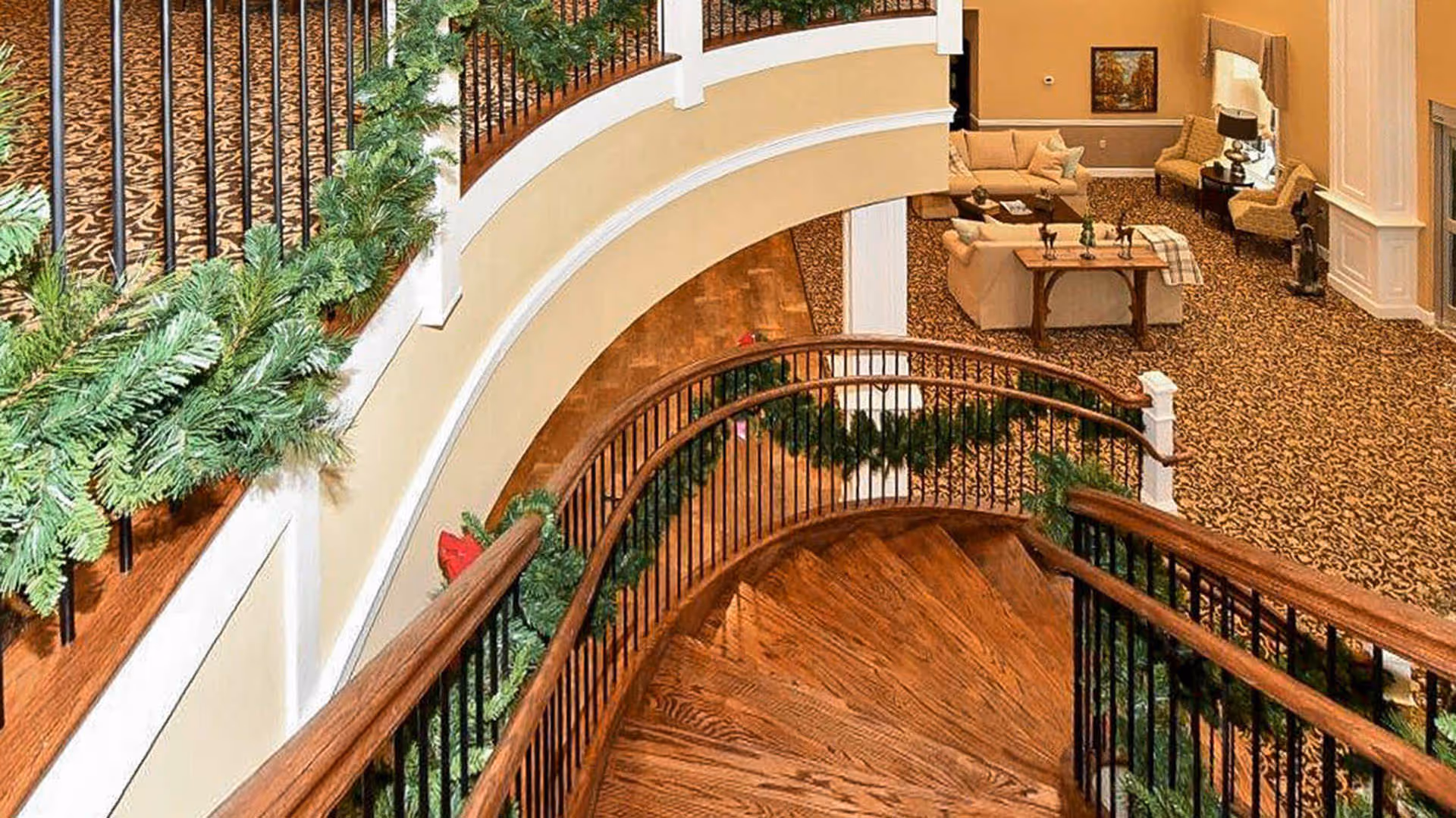 Curved wooden staircase with black metal railings decorated with green garlands, leading down to a carpeted living area with beige sofas, armchairs, a wooden coffee table, and a fireplace in a warmly lit room.