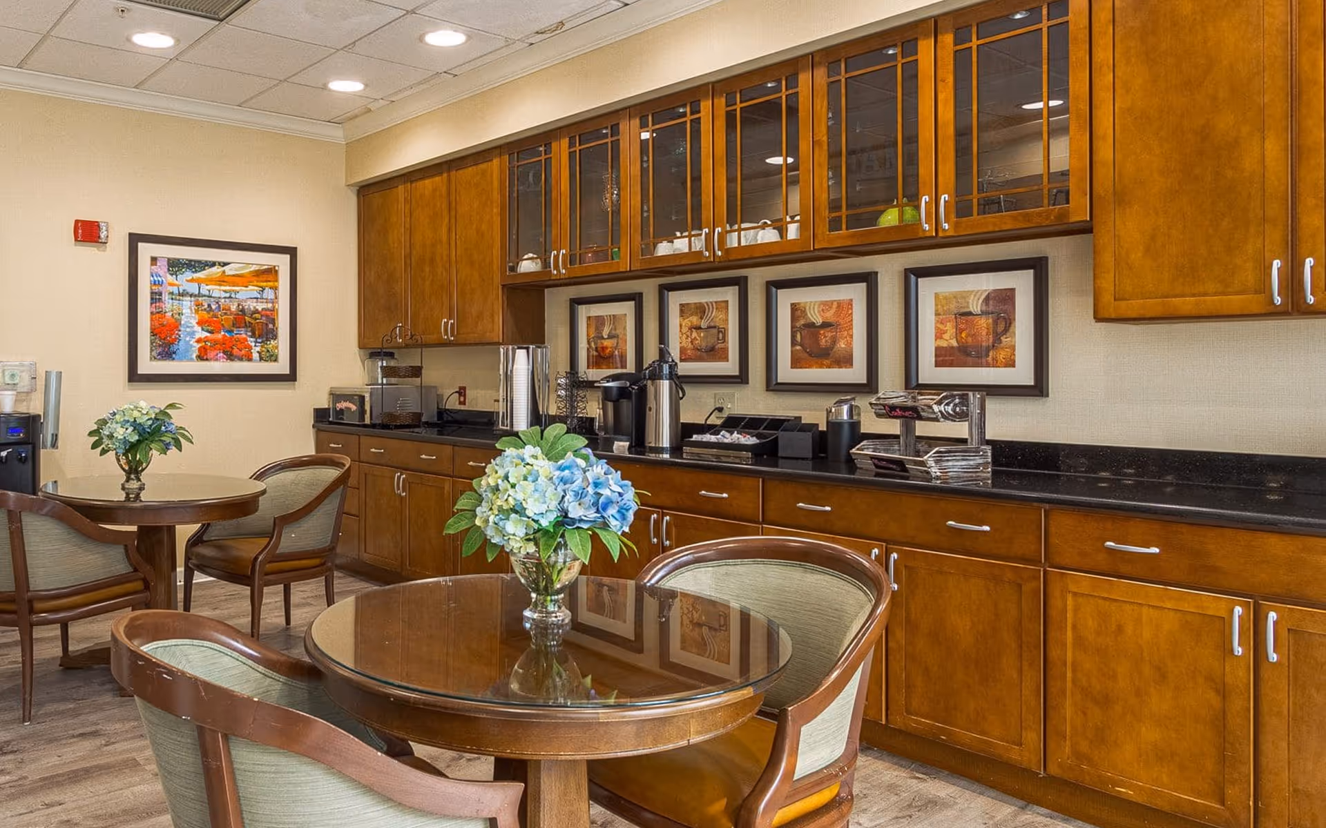 A cozy dining area with round wooden tables and cushioned chairs. Each table has a vase with blue and white flowers. The background features wooden cabinets with glass doors displaying cups and dishes, a countertop with coffee machines, and framed artwork on the wall.