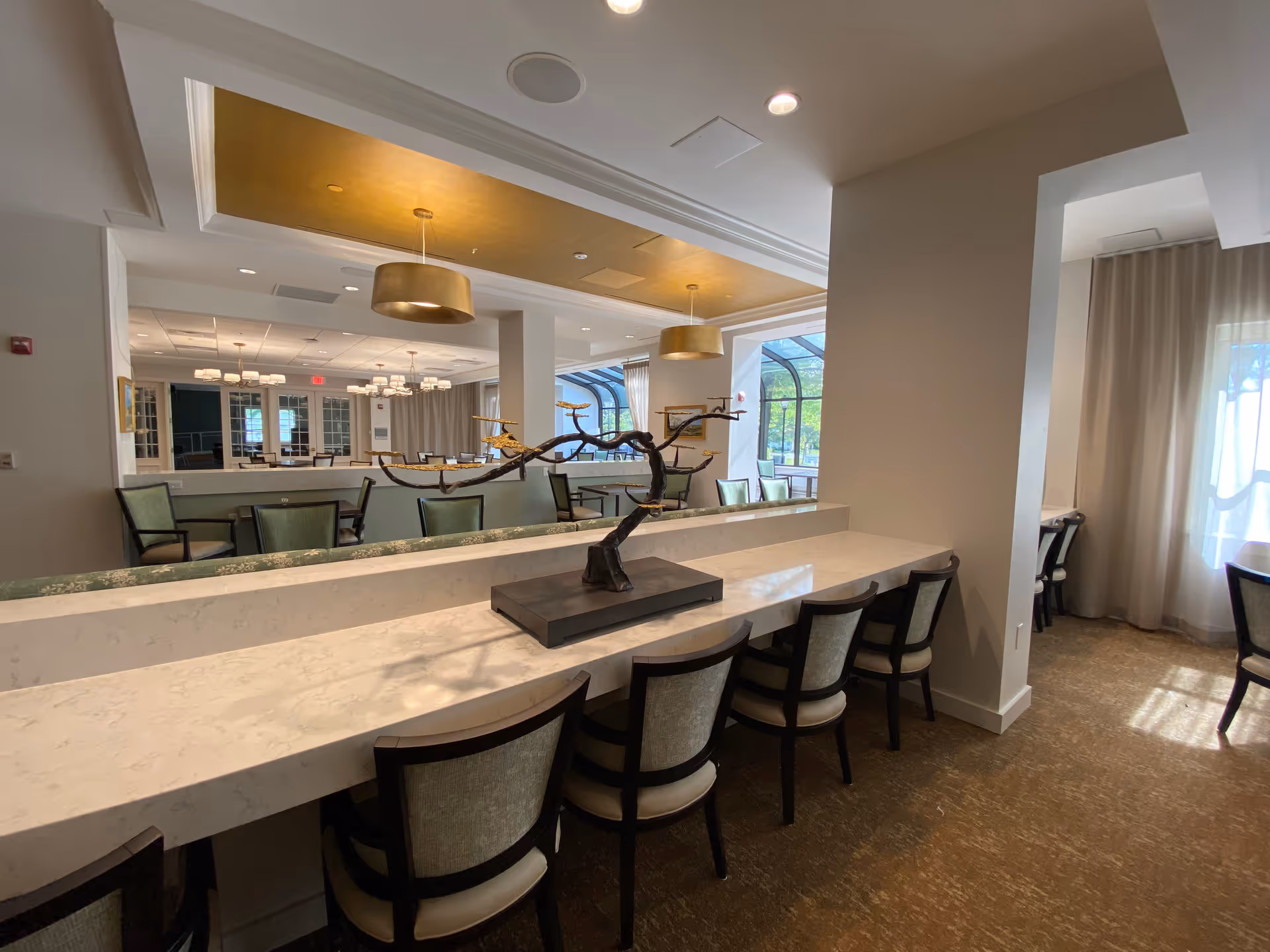 Interior view of a dining area in Westminster at Lake Ridge a CCRC, featuring a long marble countertop with several chairs lined up. A decorative sculpture resembling a tree branch with golden leaves is placed on the countertop. In the background, there are more tables and chairs, large windows with curtains, and modern hanging light fixtures with gold accents.