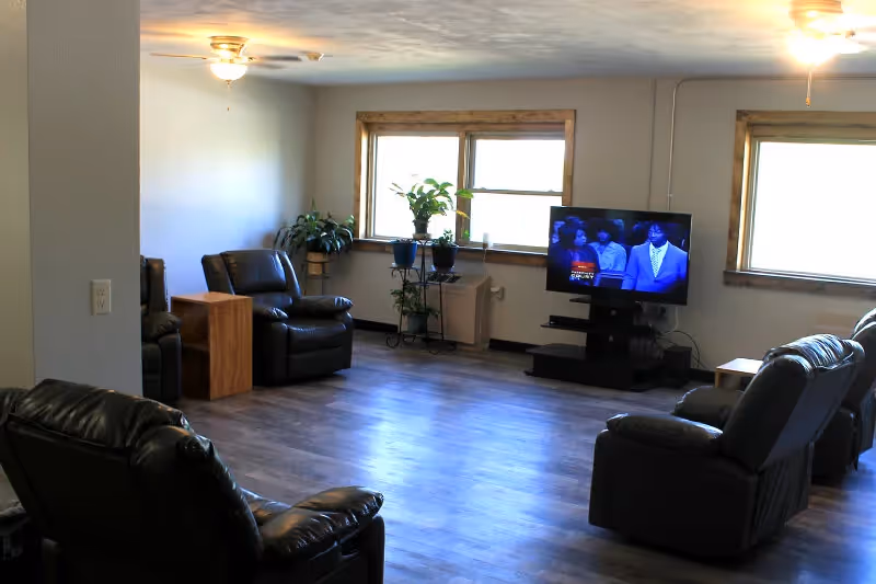 Spacious living room with black leather recliners, a TV stand, and plants by sunny windows.