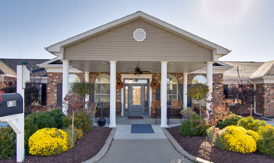 Front exterior view of Ravenwood Terrace featuring a covered entrance with white columns, brick walls, a gray door with glass panels, two wooden chairs, and landscaped garden beds with yellow flowers and shrubs on either side of the walkway.