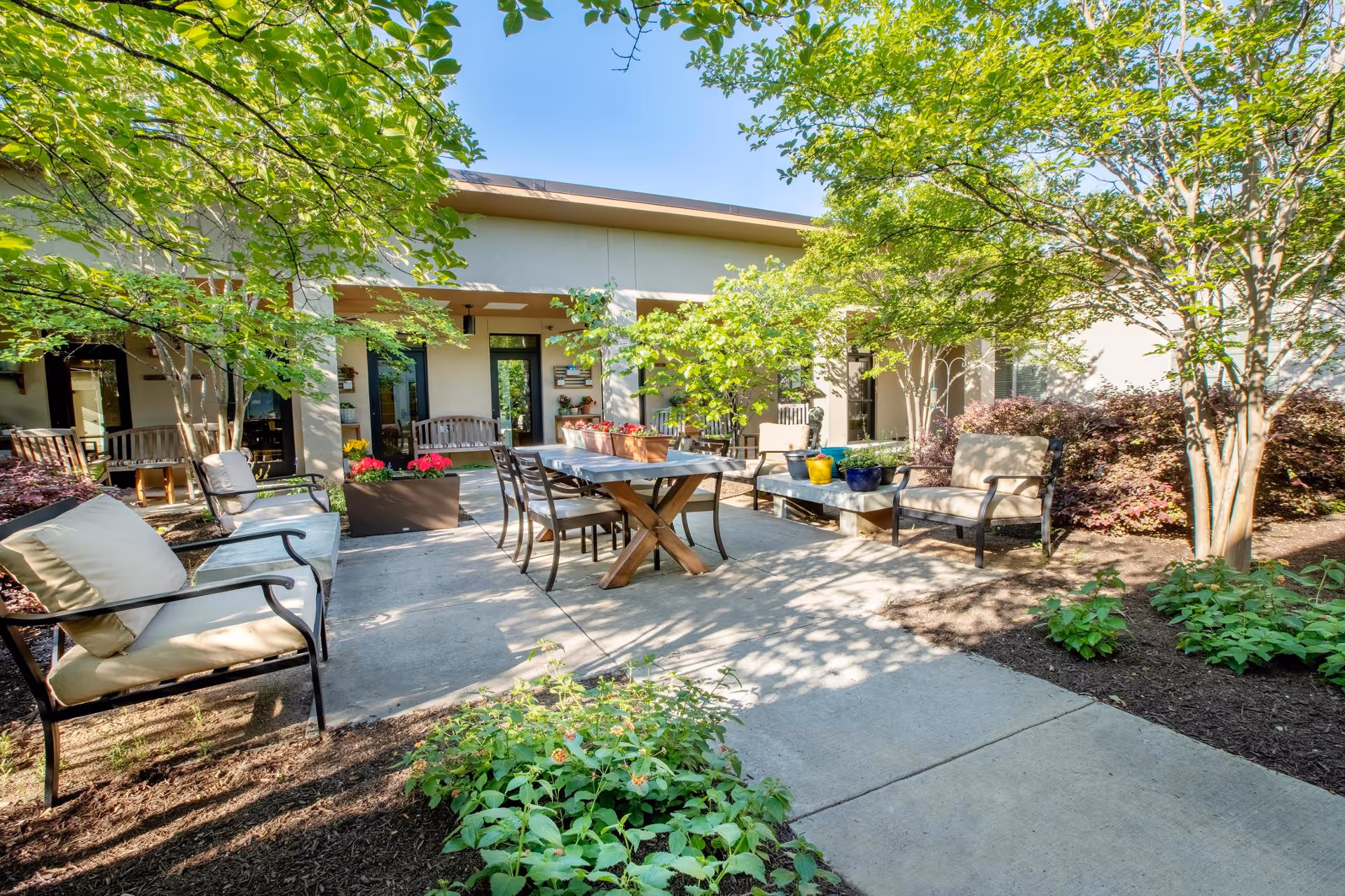 Sunlit outdoor courtyard with patio seating, a dining table, and surrounding trees and plants.