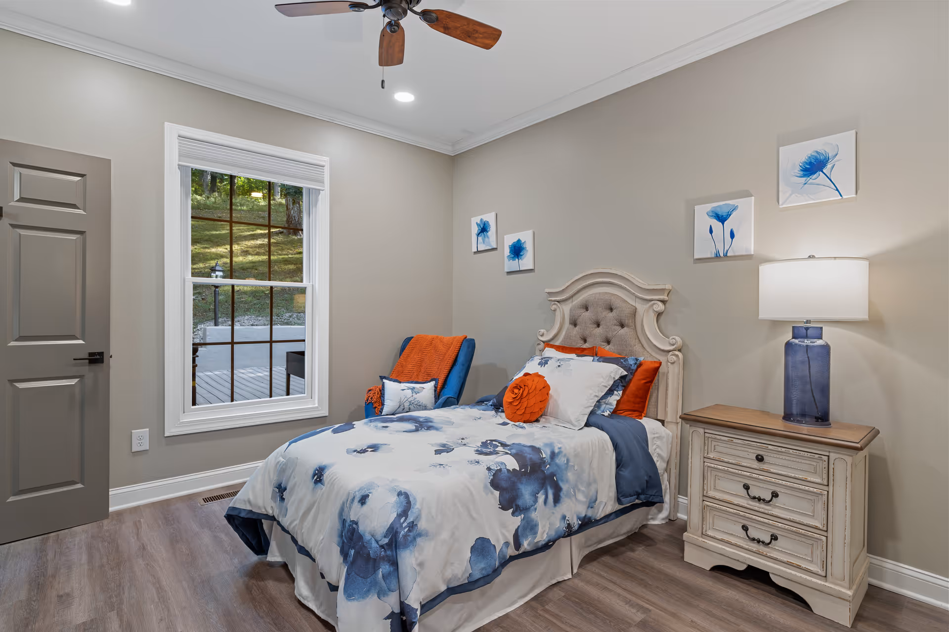 A cozy bedroom with a single bed featuring a tufted headboard and blue and white floral bedding accented with orange pillows. Next to the bed is a vintage-style nightstand with three drawers and a blue glass lamp. A blue armchair with an orange throw blanket and pillow sits in the corner near a window showing an outdoor view. The room has light beige walls, wood flooring, a ceiling fan with wooden blades, and three blue floral paintings on the wall above the bed.