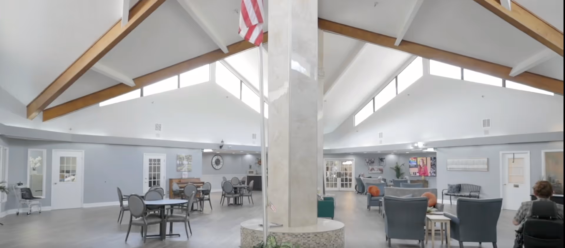 Spacious senior living facility common area with high vaulted ceilings and wooden beams. The room is divided by a central pillar with an American flag. On the left side, there are round tables with chairs arranged for dining or activities. On the right side, there is a lounge area with armchairs, a TV mounted on the wall, and a person in a wheelchair facing the TV. The walls are light gray with white doors and windows, and the floor is light wood.