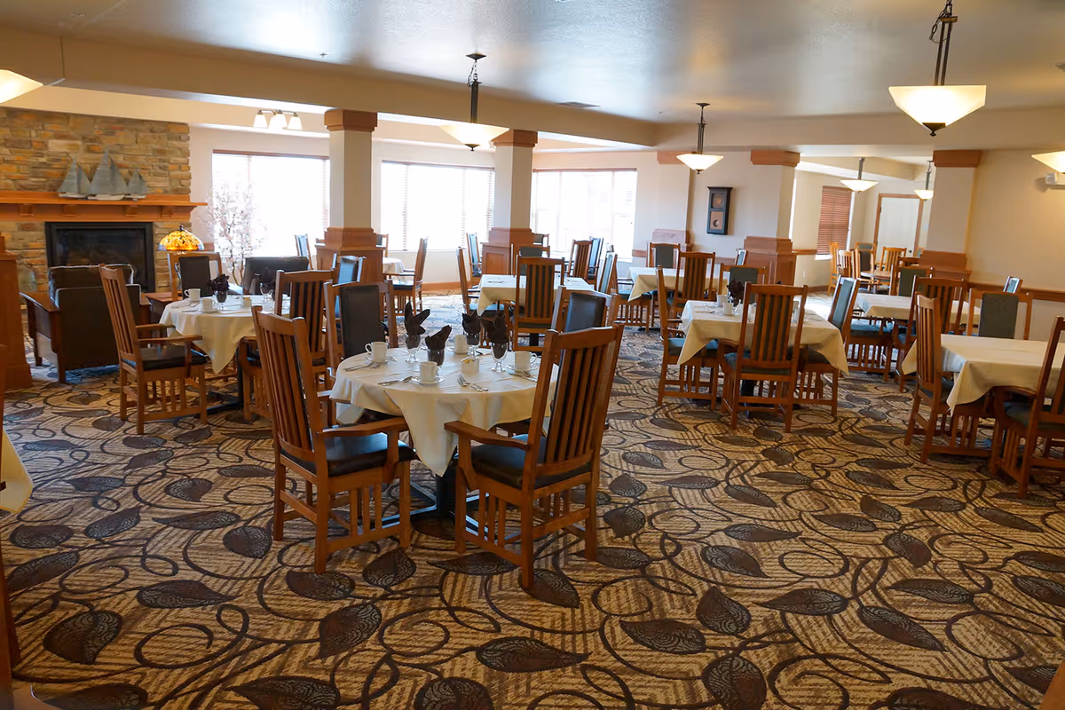 A spacious dining room with multiple wooden tables and chairs arranged neatly. Each table is covered with a white tablecloth and set with cups, glasses, and napkins. The room features a patterned carpet, large windows allowing natural light, and a stone fireplace with decorative sailboat models on the mantel.