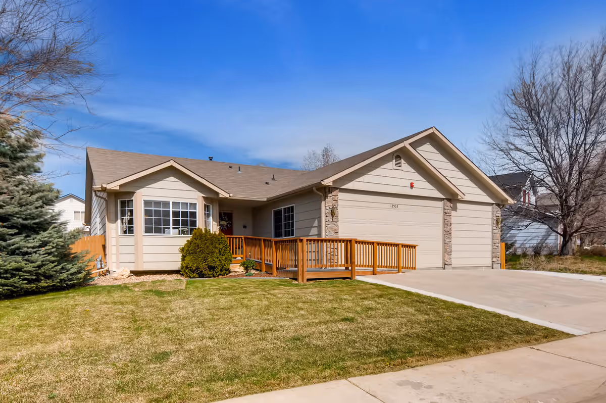 Single-story house with beige siding and a brown shingled roof, featuring a wooden wheelchair ramp leading to the front door. The house has a two-car garage and a well-maintained lawn with a few trees and shrubs under a clear blue sky.