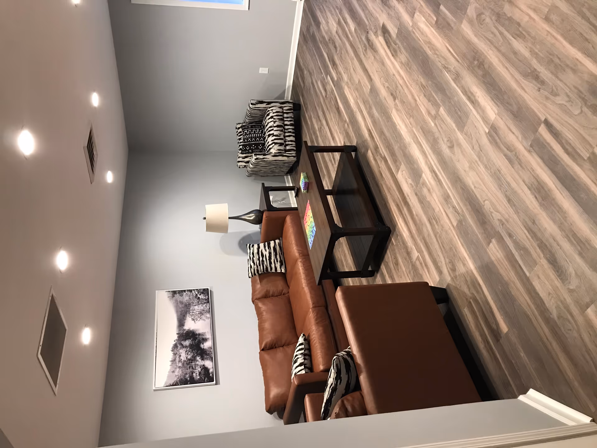 A living room with wood flooring, a brown leather sofa and matching armchair, a patterned armchair, a wooden coffee table with colorful items on it, a side table with a lamp, and a black and white landscape photograph on the wall.