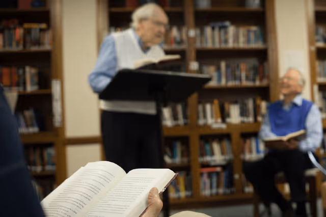 An elderly person holding an open book in the foreground with two elderly men in the background, one standing and reading from a book on a music stand, and the other sitting with a book in hand, in a room with bookshelves filled with books.
