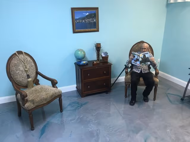 An elderly man sitting on an ornate wooden chair with upholstered seat and backrest, reading a magazine in a room with light blue walls and a polished floor. Another similar chair is empty nearby, and a small wooden cabinet with a globe, a candle holder, and other decorative items is placed against the wall. A framed picture hangs above the cabinet.